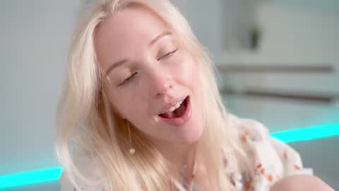 Media: Video of a smiling, fair-skinned blonde woman with long hair, wearing a floral top, eyes closed, and mouth open in a joyful expression, set against a blurred, modern kitchen background with blue LED lights.