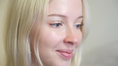 Media: Video of a close-up, smiling blonde woman with fair skin and blue eyes. Her hair is straight and shoulder-length, with a soft, natural makeup look. The background is a plain, light beige, creating a minimalistic and intimate atmosphere.