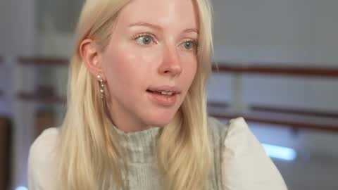 Media: Video of a fair-skinned, blonde woman with straight hair, wearing a light gray, textured top, and pearl earrings, talking with a soft expression in a modern, softly lit room with ballet barres in the background.