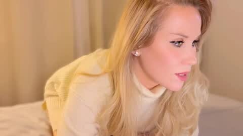 A video of a fair-skinned, blonde woman with long, wavy hair in a cream-colored sweater, leaning forward, looking pensive, in a softly lit, beige-toned room.