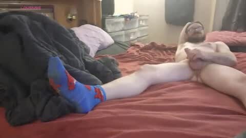 Media: Video of a nude, fair-skinned man with a beard, wearing blue socks, lying on a bed with red sheets in a cluttered, dimly lit bedroom.
