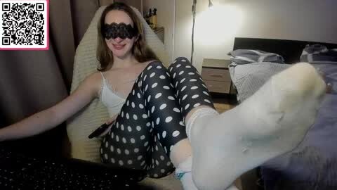 Media: A video of a young woman with fair skin and brown hair, wearing a black lace mask, a white lace bra, and black polka dot leggings, sitting on a bed in a dimly lit bedroom.