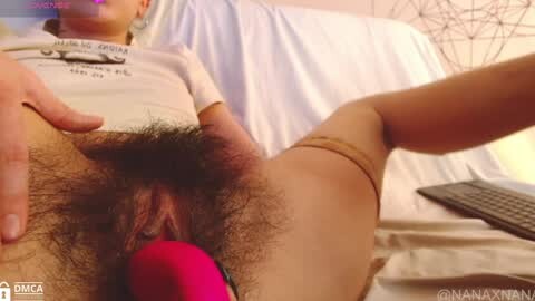 Media: A video featuring a woman with light skin and short blonde hair, lying on a bed with a white sheet, wearing a white t-shirt. Her pubic area is visible, with a pink vibrator inserted. The background includes a computer keyboard.