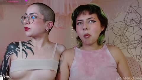 Media: Video of two women with tattoos, one with a shaved head, glasses, and red lipstick, wearing a beige bandage, the other with green hair and pink top, in a cozy room with abstract decor.