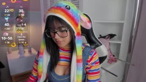 Media: Video of a young woman with glasses, rainbow unicorn hat, and striped top, posing in a bedroom with a plush unicorn, white shelves, and a pink jacket hanging.
