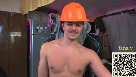Media: Video of a shirtless, fair-skinned man with a mustache, wearing an orange hard hat, standing in a cluttered room with a gaming chair and a fridge in the background.