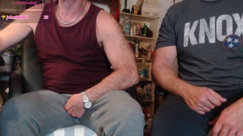 Media: Video of two men in casual attire: one in a maroon tank top, the other in a grey \"KNOW\" t-shirt, sitting side-by-side on a couch in a cluttered living room.