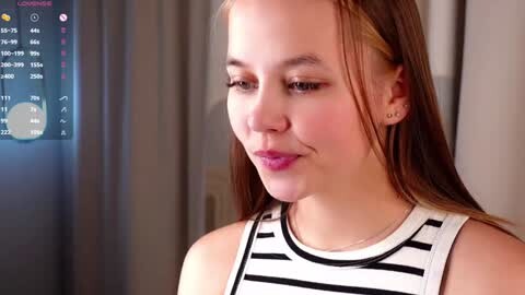 Media: Video of a fair-skinned, young woman with straight, light brown hair, wearing a black and white striped top. She stands indoors, gazing thoughtfully to the left, with a blurred background featuring a digital screen displaying a weather forecast.