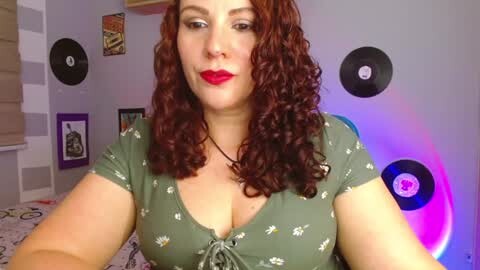 Media: A video of a curvy, fair-skinned woman with curly auburn hair, wearing red lipstick, a low-cut olive-green top with floral print, in a colorful, retro-themed room with vinyl records and framed posters on the walls.