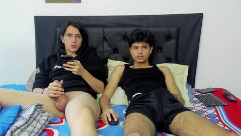 Media: Video of two young men, one with a large erect penis, lying on a bed with a colorful blanket. They wear black athletic shorts and hoodies.