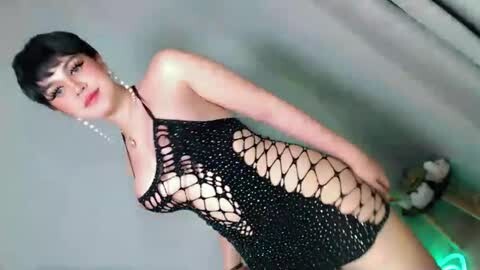 Media: Video of an Asian woman with short black hair and pale skin, wearing a revealing black fishnet bodysuit that exposes her breasts and stomach, standing in a dimly lit room with a blurred background.