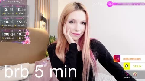 Media: A video of a fair-skinned woman with long, light brown hair, wearing a black top, smiling at the camera. She's seated on a bed with a floral pillow in a modern, light-colored bedroom. A virtual chat window is overlaid on the image.