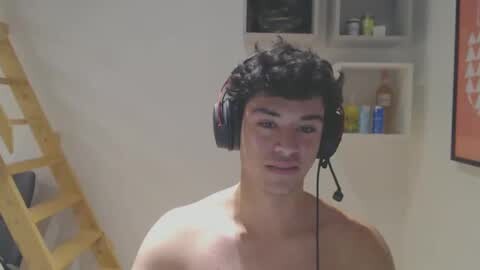 Media: A video of a shirtless young man with curly black hair wearing a headset, standing in a simple, cluttered room with a wooden ladder and shelves holding toiletries.