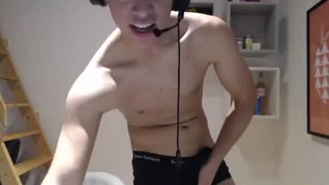 Media: Video of a shirtless, fit young man with short brown hair, wearing black underwear, speaking into a headset microphone, standing in a modern, minimalist room with beige walls and a wooden ladder.