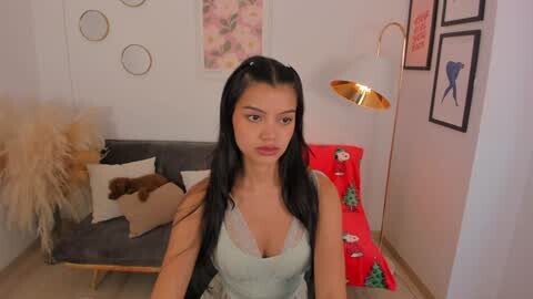 Media: A video of a young woman with long black hair and light skin, wearing a light blue tank top, standing in a cozy living room with beige pillows, a Christmas-themed throw, and framed artwork.