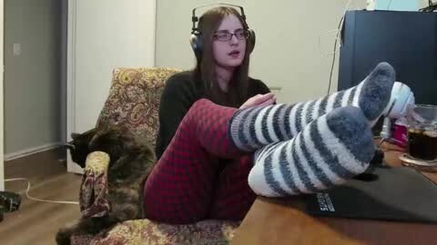 Media: Video of a woman with long brown hair, wearing glasses and headphones, sitting in a patterned armchair, legs up in striped socks, surrounded by a cluttered room.