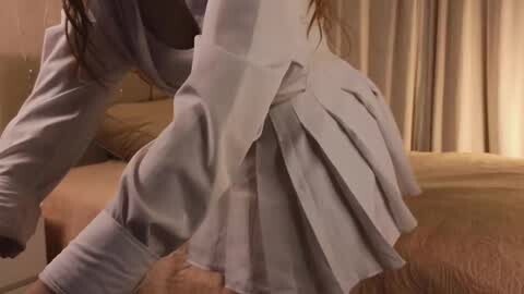 Media: Video of a person in a grey pleated skirt and white shirt bending over, in a beige bedroom with cream curtains and a bed with a beige blanket.