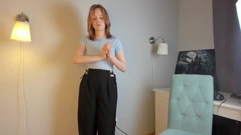 Media: Video of a slim, light-skinned woman with shoulder-length red hair in a light blue shirt and high-waisted black pants, standing in a modern, minimalistic room with a teal chair, gray walls, and a framed video.