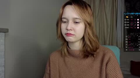 Media: A video of a young woman with shoulder-length, light brown hair, wearing a beige ribbed sweater, seated in a dimly lit room with a computer screen displaying a video game interface.