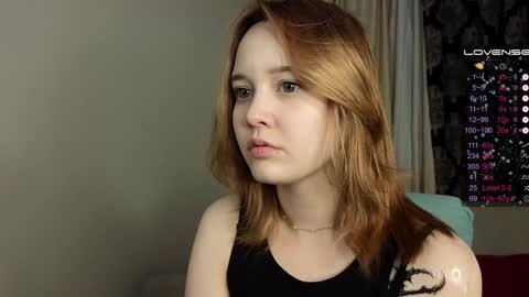 Media: A video of a fair-skinned, red-haired young woman with a serious expression, wearing a black top, against a dimly lit background with a calendar and abstract artwork.