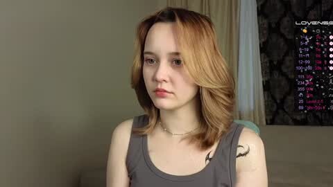 Media: Video of a young Caucasian woman with light skin and shoulder-length, wavy auburn hair, wearing a gray tank top, sitting indoors against a beige wall with a black and white wall calendar in the background.