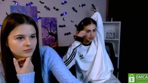 Media: Video of two young women with pale skin, one with long dark hair, the other with medium-length dark hair, in a cozy room decorated with bat-themed wall art and glowing blue lights.
