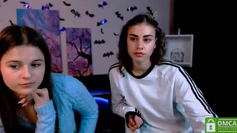 Media: Video of two teenage girls with fair skin and dark hair; one in a blue sweater, the other in a white hoodie, indoors with Halloween decorations and bat posters.