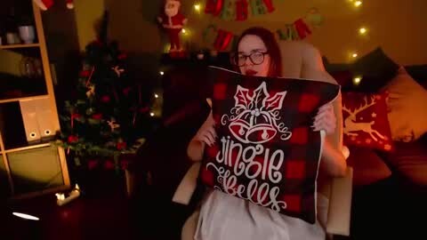 Media: Video of a woman in glasses, red hair, and a white dress, sitting on a beige chair, holding a black and red plaid pillow with \"Jingle Bell Blues\" text. Background includes a lit Christmas tree, a Santa toy, and festive decorations.