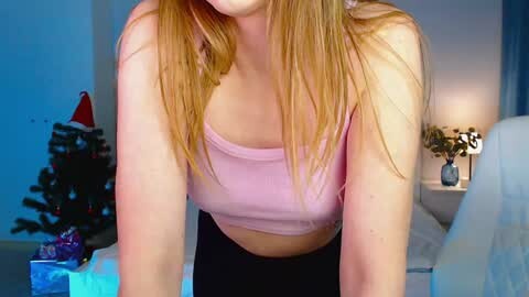Media: Video of a slender, light-skinned woman with long, straight blonde hair, wearing a pink tank top and black pants, leaning forward, indoors, near a decorated Christmas tree.