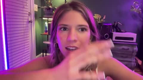 Media: Video of a young woman with light skin and brown hair, wearing minimal makeup, sitting in a dimly lit room with purple lighting. She is smiling and making a peace sign with her fingers.