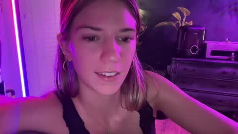 Media: Video of a young woman with light skin, straight shoulder-length blonde hair, wearing a black lace top. She's in a dimly lit room with purple lighting, holding a phone for a selfie.