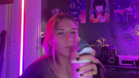 Media: A video of a young woman with light skin, straight brown hair, and a small tattoo, holding a drink in a dimly lit room with neon lights and anime posters.