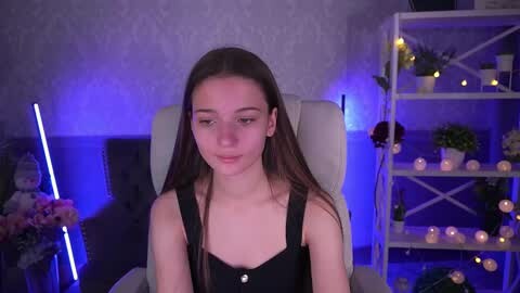 Media: Video of a young, slender, Caucasian girl with long brown hair, wearing a black tank top, sitting on a white chair in a dimly lit, cozy room with a blue glow, bookshelves, and candles.