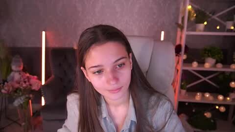 Media: Video of a young woman with straight brown hair, wearing a light blue denim shirt, seated in a modern, dimly lit room with white furniture, floral arrangements, and string lights in the background.
