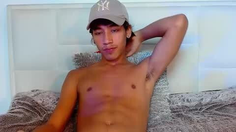 Media: Video of a shirtless, young Hispanic male with short hair, wearing a New York Yankees cap, lying on a bed with a gray fur blanket and white headboard.