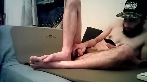 Media: A video of a bearded, nude man with pale skin, wearing a baseball cap, playing a video game on a laptop, lying on a couch.