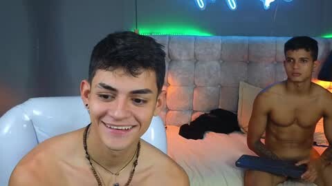 Media: Video of two shirtless young men in a dimly lit bedroom with a white leather chair and a tufted headboard. One man has short dark hair, a gold chain, and a tattoo on his chest; the other, with a muscular build, holds a blue remote.