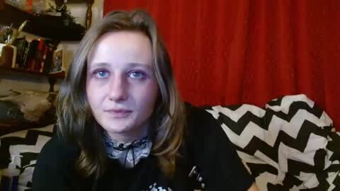 Media: Video of a young Caucasian woman with fair skin and light brown hair, sitting on a black and white zebra-patterned couch. She wears a black top and a grey scarf. Background features a red curtain and a messy shelf with various items.
