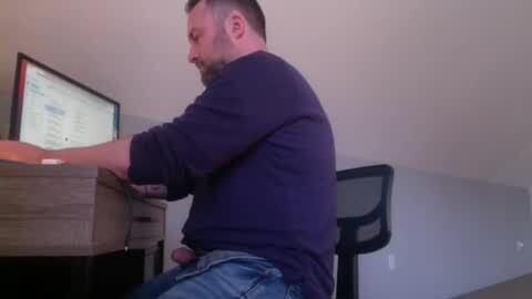 Media: Video of a bearded, short-haired white man with a medium build, wearing a dark blue shirt and jeans, sitting at a wooden desk in a minimalist office.