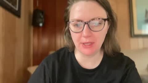 Media: Video of a fair-skinned woman with shoulder-length brown hair and glasses, wearing a black shirt, sitting in a warmly lit room with wooden walls and a framed painting in the background.