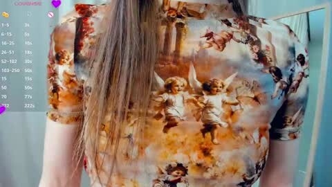 Media: A video of a woman with long brown hair wearing a short-sleeved, long-sleeved, patterned top depicting classical angels in motion against a fiery background. She stands indoors, partially obscured by a calendar and blurred background.