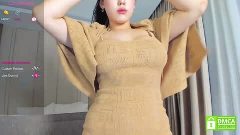 Media: A video of an East Asian woman with fair skin, wearing a beige, ribbed dress, and a matching open cardigan, with her arms raised, against a modern, light-colored background.