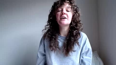Media: Video of a young girl with long, curly brown hair, wearing a light blue shirt, eyes closed, smiling, in a softly lit room with white walls.
