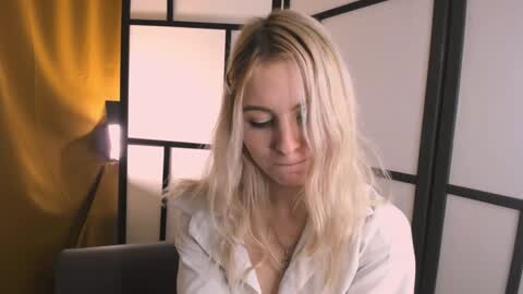 Media: Video of a young, fair-skinned woman with long, straight blonde hair, wearing a white blouse, standing in front of a shoji screen. Background includes a yellow curtain and a modern lamp.