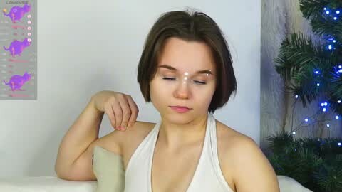 Media: Video of a young woman with short brown hair and fair skin, wearing a white halter top, applying makeup. She sits on a white couch, with a map and a potted plant in the background.