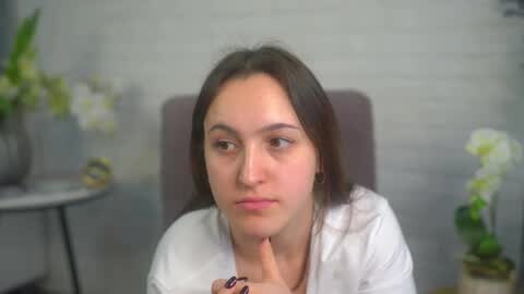Media: Video of a young Caucasian woman with fair skin and long, straight brown hair, wearing a white blouse, sitting in a chair in a modern room with grey walls and a white orchid in a pot on the left.