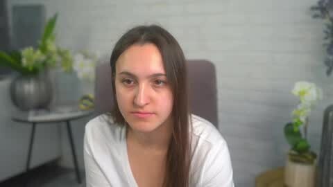 Media: Video of a young woman with long brown hair, wearing a white robe, sitting indoors with a neutral expression. Background features a white brick wall, potted plants, and a vase with white flowers.