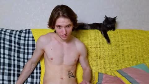 Media: Video of a shirtless, slim, young man with shoulder-length brown hair and a small tattoo on his left side, sitting on a yellow sofa, with a black and white checkered blanket, and a gray cat resting on his shoulder.