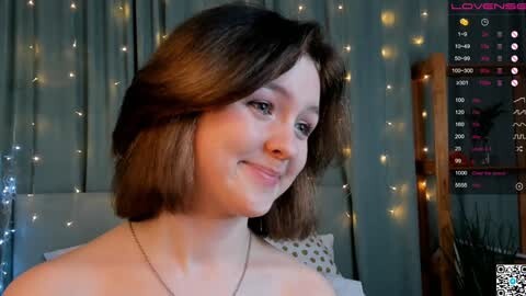 Media: Video of a young woman with short, straight brown hair, fair skin, and a subtle smile, wearing a delicate necklace. She stands against a softly lit backdrop with string lights and a wooden ladder, and a digital screen showing live stream details.