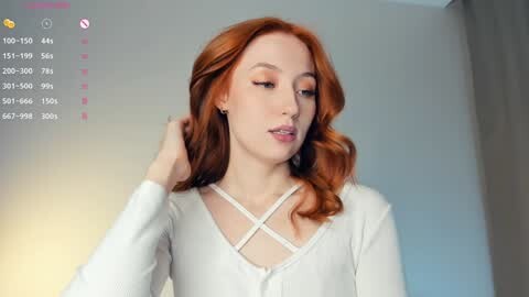 Media: Video of a fair-skinned, red-haired woman with wavy hair, wearing a white cardigan with a crisscross neckline, set against a soft-focus background.